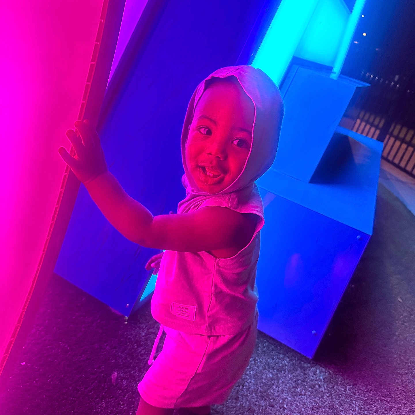 Takari is registered to the contest to win money with this photo: barefoot, blue_light, child, clothing, colorful, curious, glowing, hood, illumination, joyful, neon_lights, night, outdoor, person, pink_light, playful, smile, standing, texture, toddler