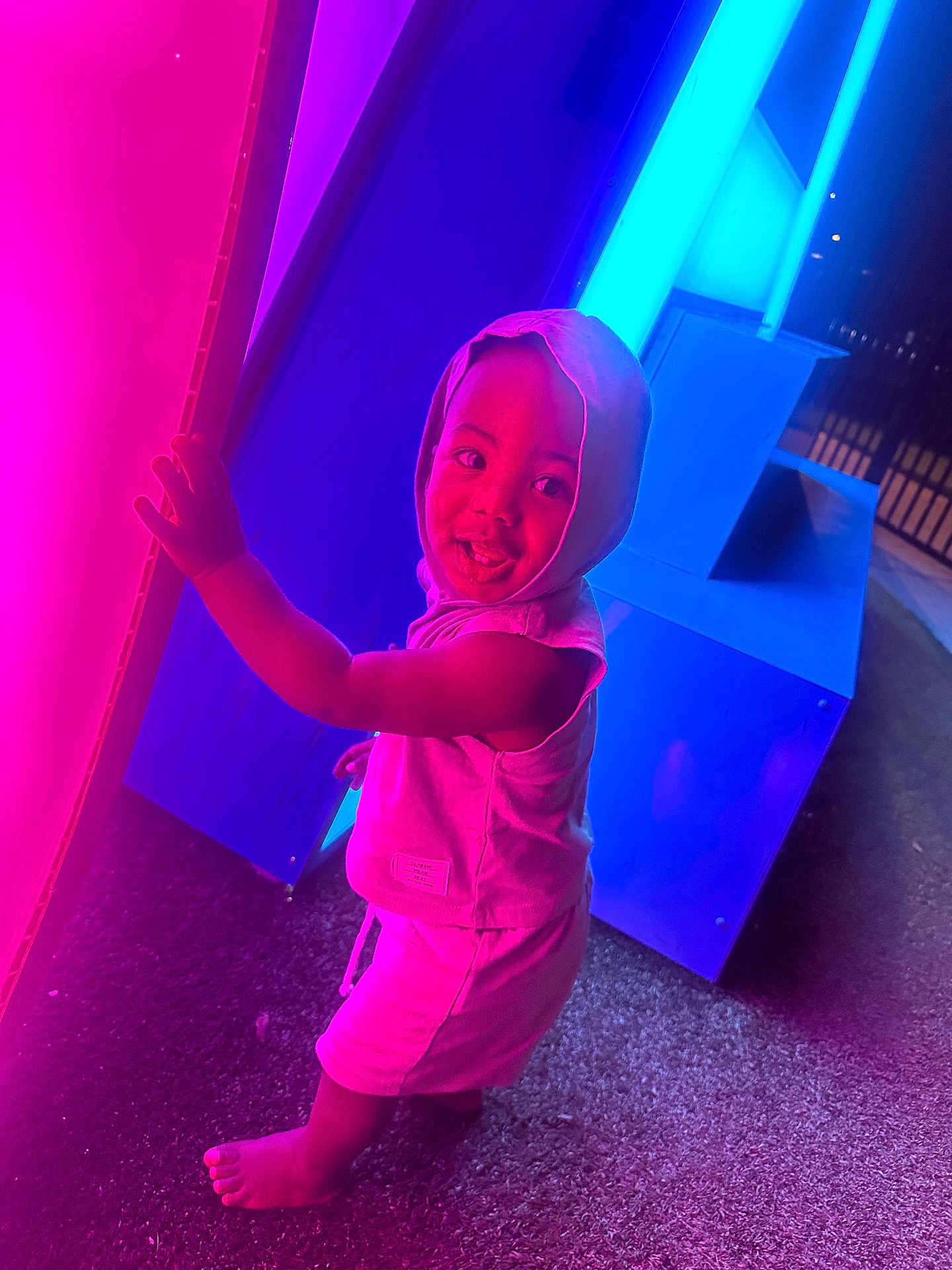 Takari is registered to the contest to win money with this photo: child, toddler, hood, smile, barefoot, neon_lights, pink_light, blue_light, night, glowing, curious, standing, outdoor, playful, colorful, person, clothing, texture, joyful, illumination