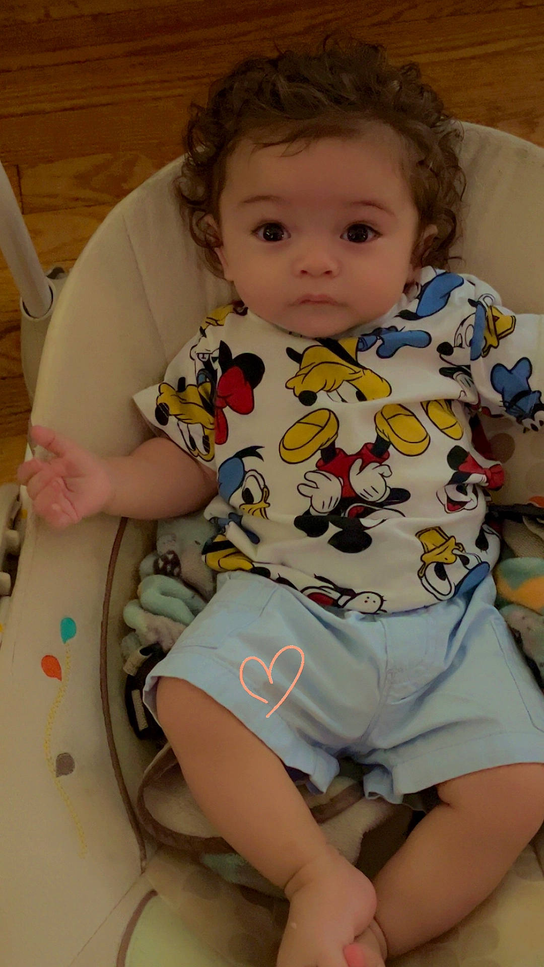 adorable, baby, baby_seat, cartoon_shirt, child, clothing, comfort, curly_hair, cushion, cute, hand_drawn, indoor, infant, light_shorts, looking_at_camera, person, portrait, sitting, small_heart, wooden_floor