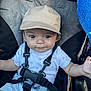 baby, child, cap, stroller, seatbelt, clothing, shorts, white_shirt, curly_hair, outdoor, sunlight, face, hands, safety_harness, infant, cute, casual, expression, seat, summer