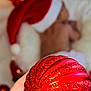 child, santa_hat, red_ornament, hand, holiday_lights, christmas, blurred_background, festive, decoration, glitter, sleeping, indoors, celebration, soft_focus, holiday_season, festive_colors, cozy, person, closeup, warm_lighting
