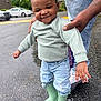 adult_hand, casual, child, cute, fashion, green_boots, jeans, outdoor, pavement, person, playful, rain_boots, rainy_day, smile, standing, street, support, sweater, toddler, wet_surface