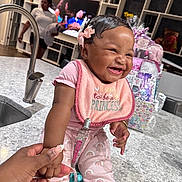 Mylah joined the competition — help win amazing prizes! baby, smiling, laughing, pink_clothing, bib, hand_holding, kitchen, sink, faucet, adult_hand, woman, television, modern_interior, countertop, gift_bags, hair_bow, happy, child, indoor, portrait