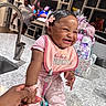 baby, smiling, laughing, pink_clothing, bib, hand_holding, kitchen, sink, faucet, adult_hand, woman, television, modern_interior, countertop, gift_bags, hair_bow, happy, child, indoor, portrait