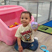 Prince is registered to the contest to win money with this photo: toddler, child, toy, playroom, pink_toy_bin, colorful_balls, floor, wooden_floor, fence, indoor, smile, casual_clothing, blue_jeans, white_shoes, short_sleeve_shirt, curly_hair, holding_toy, play_area, plastic_toy, cute