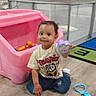 toddler, child, toy, playroom, pink_toy_bin, colorful_balls, floor, wooden_floor, fence, indoor, smile, casual_clothing, blue_jeans, white_shoes, short_sleeve_shirt, curly_hair, holding_toy, play_area, plastic_toy, cute