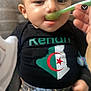 Kenan a rejoint le concours — aidez-le/la à gagner de superbes lots ! baby, child, feeding, spoon, food, black_shirt, algerian_flag, cushion, striped_background, hand, clothing, smile, face, indoor, cute, infant, person, sitting, apparel, young