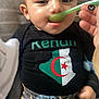 algerian_flag, apparel, baby, black_shirt, child, closeup, cushion, cute, feeding, food, hand, indoor, infant, pants, person, portrait, relaxed, spoon, stars_pattern, striped_background