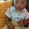 baby, blue_shirt, bow_tie, boy, child, couch, curious_expression, eyes, face, gold_bracelet, gold_chain, indoors, mobile_screen, photography, pillow, portrait, sitting, suspenders, user_interface, yellow_cushion
