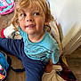 child, blonde_hair, blue_shirt, pants, sitting, blanket, floor, wooden_floor, backpack, toys, smile, face, curly_hair, indoor, casual_clothing, young, playful, messy_face, cozy, relaxed