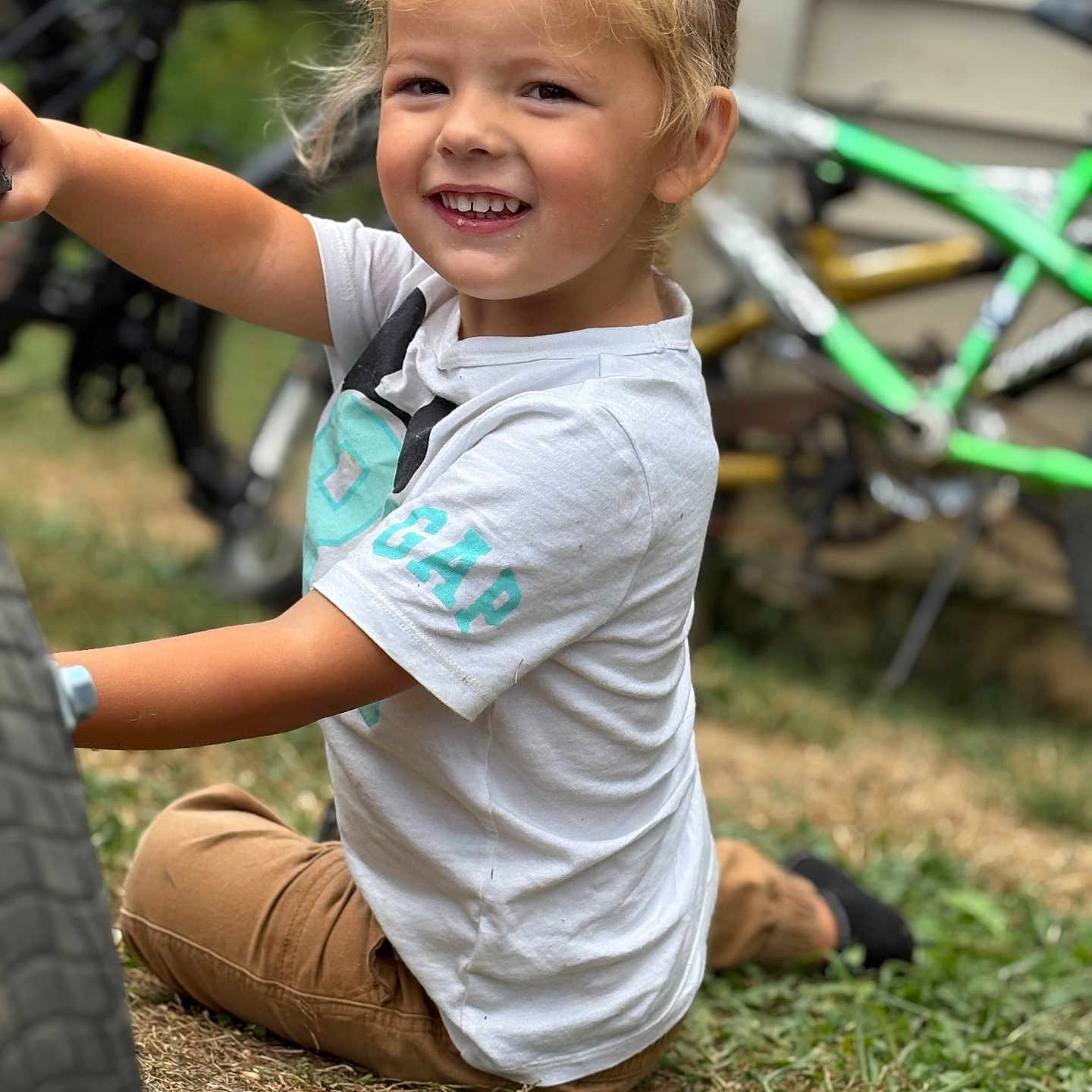 Raylon joined the competition — help win amazing prizes! bicycle, blonde_hair, brown_pants, casual_clothing, child, daylight, fun, grass, happy, nature, outdoor, person, playful, sitting, smiling, toddler, toy, wheel, white_shirt, young