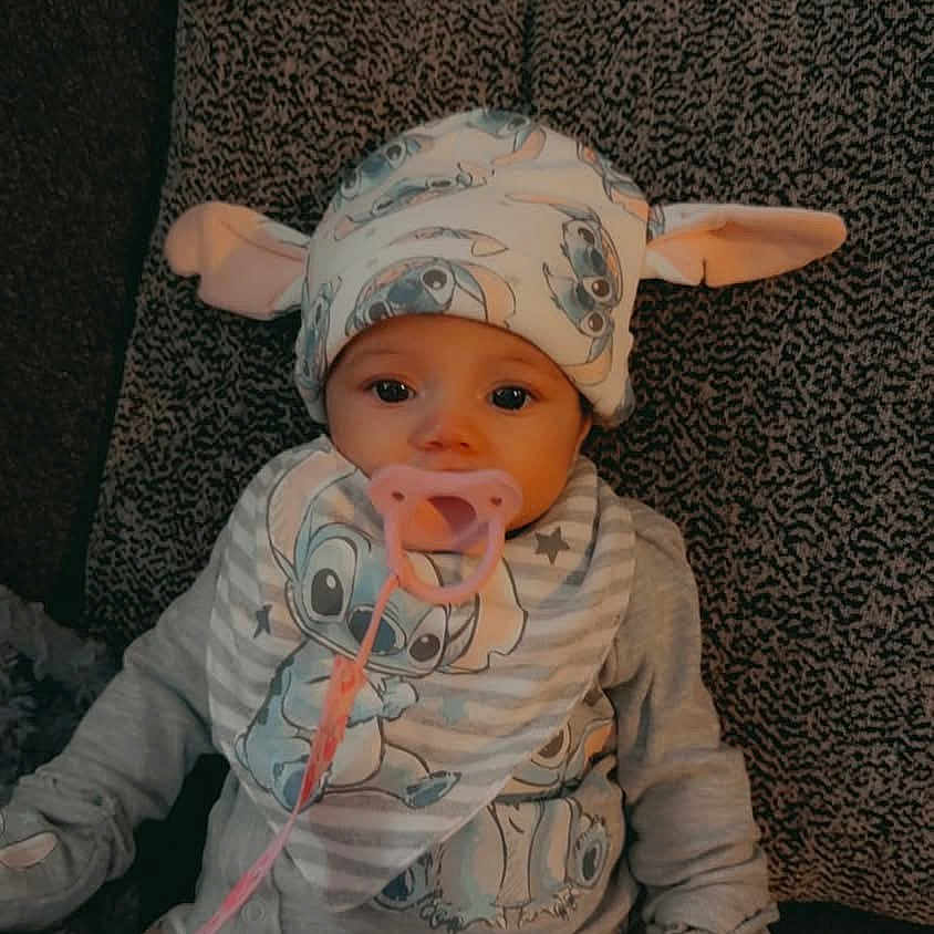 Xaviana is registered to the contest to win money with this photo: baby, child, couch, cozy, cushion, cute, expressionless, face, gray, hat, indoors, infant, pacifier, pajamas, patterned_clothing, pink, sitting, soft_lighting, stitch_character, warm