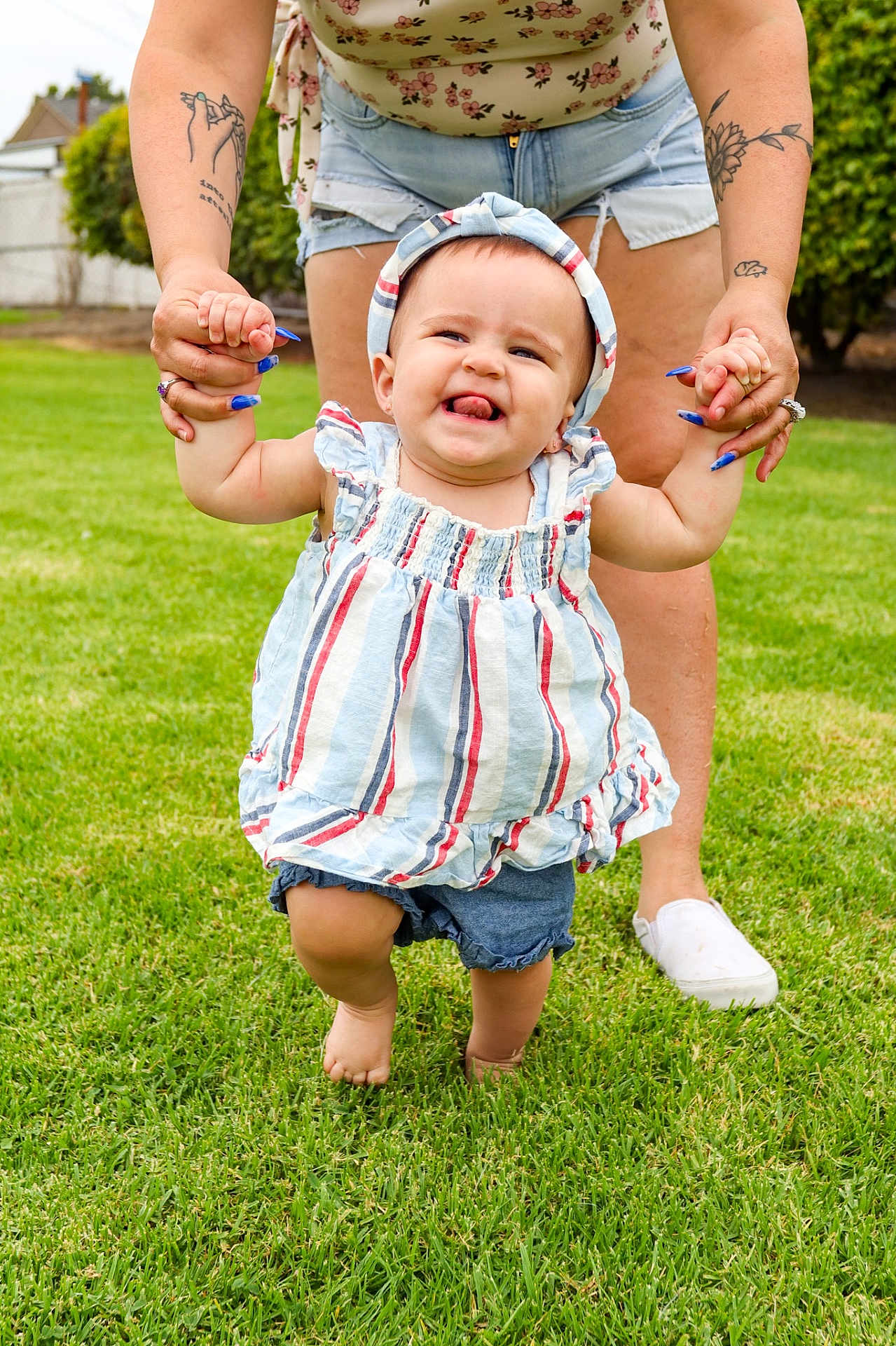 Octavia is registered to the contest to win money with this photo: toddler, child, baby, grass, outdoor, walking, holding_hands, adult, tattoo, shorts, headband, striped_dress, barefoot, nails, person, summer, smile, happy, learning_to_walk, family