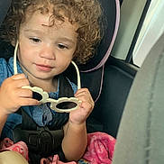 Myrakle is registered to the contest to win money with this photo: toddler, child, car_seat, curly_hair, sunglasses, pacifier, blanket, seatbelt, indoor, vehicle, baby, cute, portrait, hands, pink, blue_clothing, safety, infant, car_interior, focus