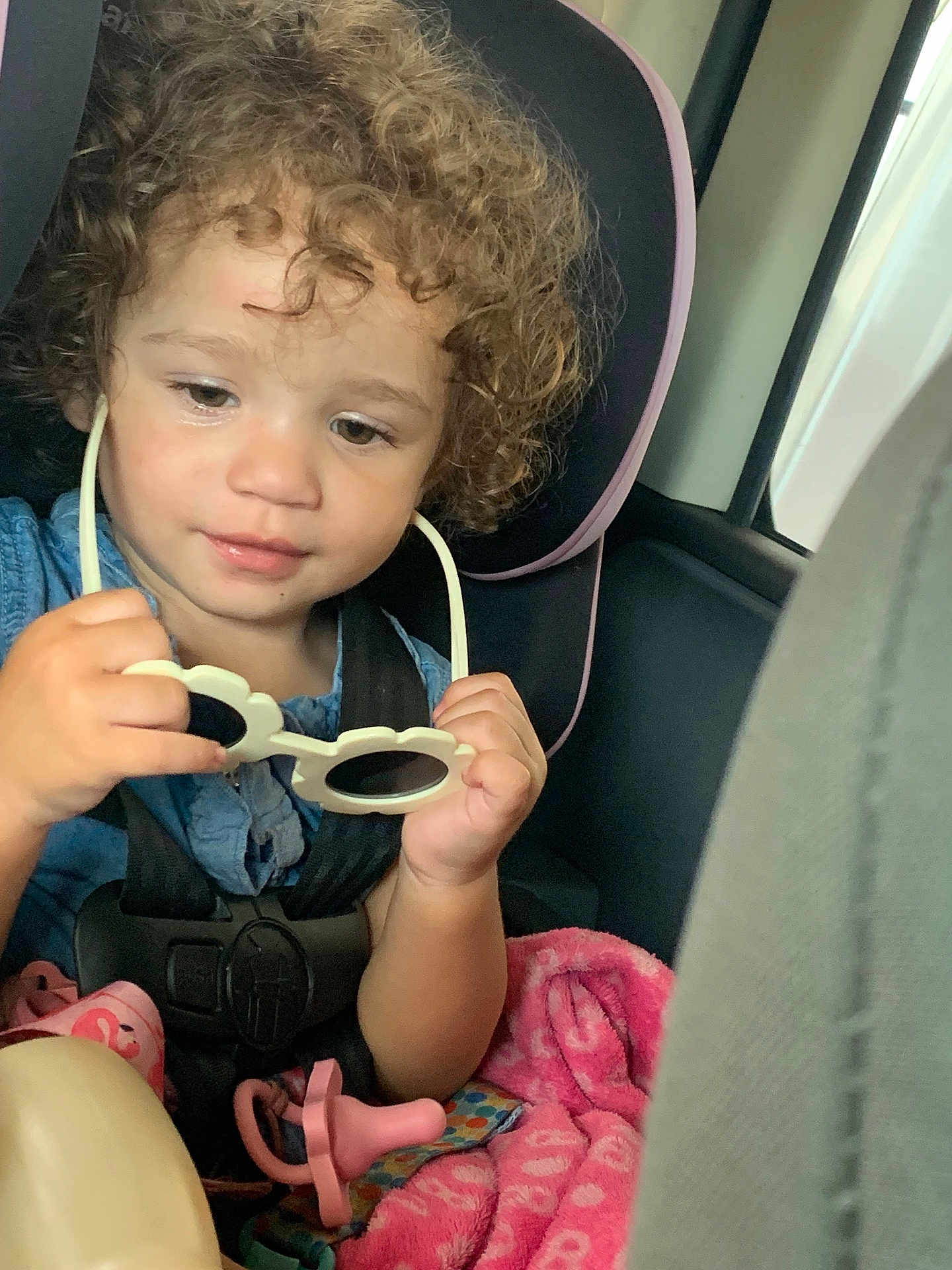 Myrakle is registered to the contest to win money with this photo: toddler, child, car_seat, curly_hair, sunglasses, pacifier, blanket, seatbelt, indoor, vehicle, baby, cute, portrait, hands, pink, blue_clothing, safety, infant, car_interior, focus