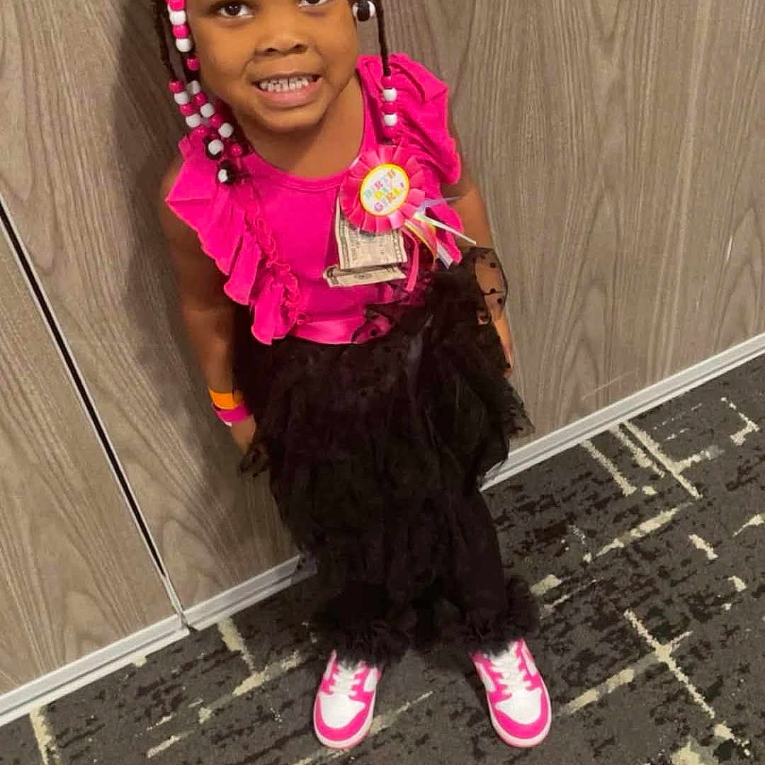 Nakhya Embry is registered to the contest to win money with this photo: badge, beads, birthday, black_skirt, braids, carpet, celebration, child, fashion, girl, happy, indoor, money, person, pink_shirt, ribbons, smiling, sneakers, standing, wall