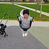 baby, child, swing, playground, outdoor, smiling, winter_clothing, hat, jacket, shoes, grass, sidewalk, stroller, suburban, street, trees, houses, playground_surface, portrait, daylight