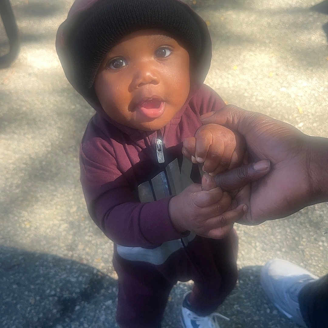 Keyon is registered to the contest to win money with this photo: baby, child, infant, portrait, outdoor, standing, beanie, winter_clothing, jacket, maroon_clothing, sneakers, adult_hand, holding_finger, playground, pavement, sunlight, shadows, serious_expression, looking_at_camera, full_body