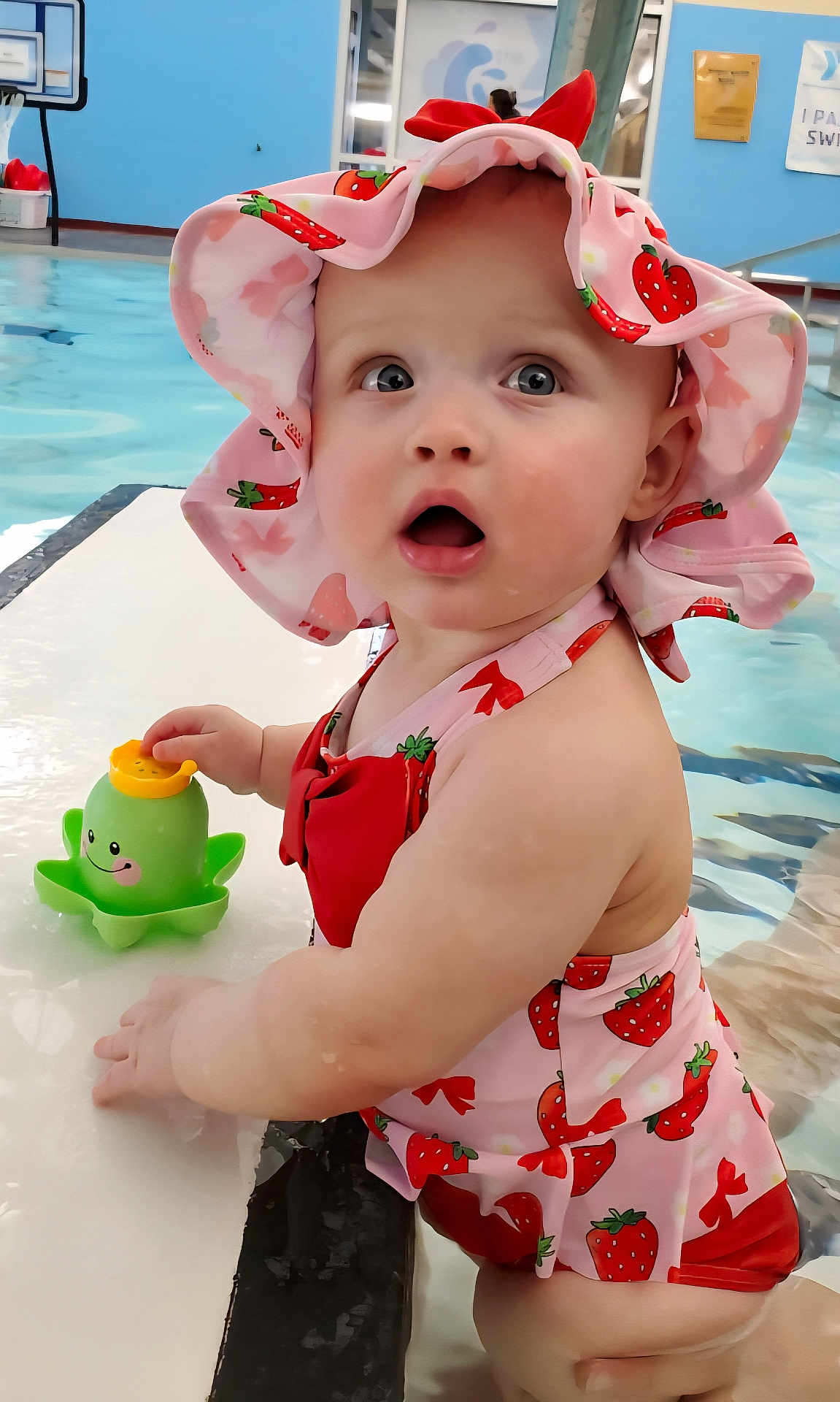 Esmae is registered to the contest to win money with this photo: baby, child, swimsuit, hat, strawberry_pattern, pool, water, toy, octopus_toy, curious, expression, summer, outdoor, playful, cute, skin, pink, red, childhood, fun