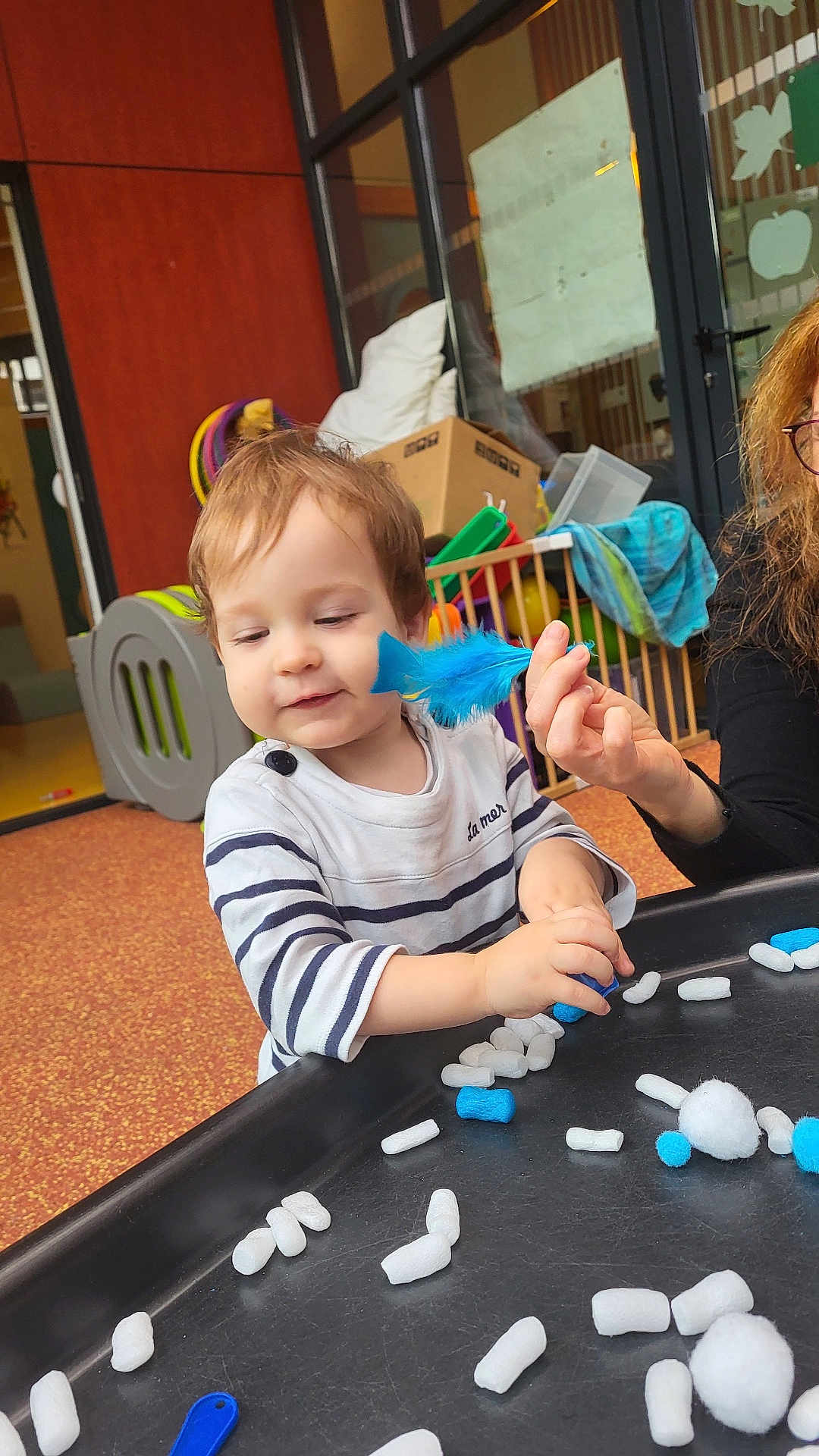 Ethan a rejoint le concours — aidez-le/la à gagner de superbes lots ! toddler, child, indoor, play, crafts, blue_feather, striped_shirt, table, toys, person, hand, face, curious, activity, colorful, playroom, window, box, floor, adult_hand