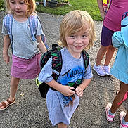 Thomas is registered to the contest to win money with this photo: child, children, backpack, outdoor, pavement, grass, house, mailbox, smile, blonde_hair, casual_clothing, blue_shoes, purple_skirt, sneakers, sunlight, playground, friends, summer, young, happy