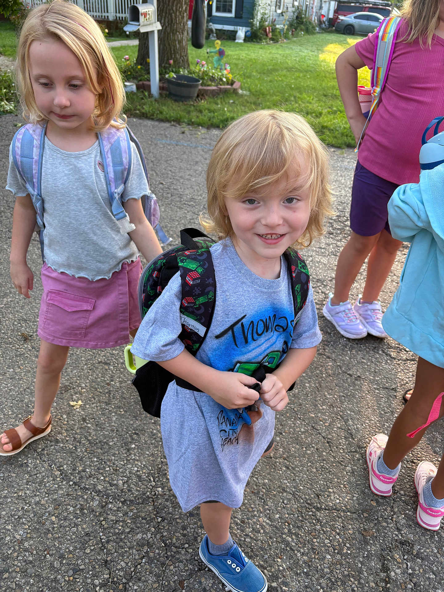 Thomas is registered to the contest to win money with this photo: child, children, backpack, outdoor, pavement, grass, house, mailbox, smile, blonde_hair, casual_clothing, blue_shoes, purple_skirt, sneakers, sunlight, playground, friends, summer, young, happy