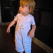 Lissandro a rejoint le concours — aidez-le/la à gagner de superbes lots ! acoustic_foam, barefoot, blond_hair, casual_clothing, child, curious, dark_background, expression, floor, indoor, indoors, looking_away, person, portrait, short_sleeves, soft_lighting, standing, toddler, white_overalls, young_child