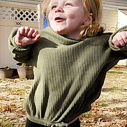 Ember is registered to the contest to win money with this photo: casual_clothing, cheerful, child, daylight, face, fence, fun, grass, green_sweater, happy, nature, outdoor, person, playful_pose, potted_plants, smile, sunlight, tree_shadow, yard, young