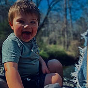 Axell is registered to the contest to win money with this photo: child, toddler, boy, portrait, smiling, tongue_out, sitting, outdoors, nature, forest, trees, blanket, clothing, shorts, t_shirt, happy, face, bokeh, sunlight, photography
