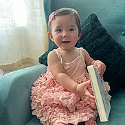 Elliana joined the competition — help win amazing prizes! baby, child, girl, pink_dress, armchair, book, smiling, cute, indoors, curly_hair, hair_clip, barefoot, portrait, happy, soft_light, cushion, seated, curtains, room, cozy