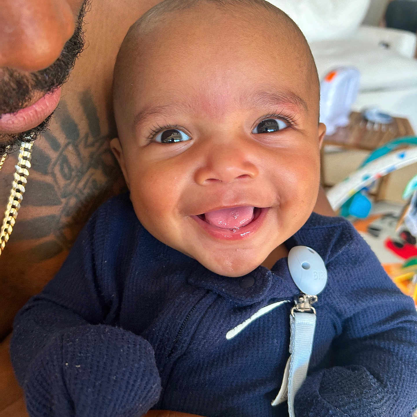 Asher is registered to the contest to win money with this photo: adult_hand, baby, blanket, blue_clothing, child, closeup, cute, face, furniture, happy, holding, home, indoor, infant, living_room, person, portrait, smiling, tattoo, tongue