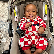 Asher is registered to the contest to win money with this photo: baby, smiling, child, stroller, onesie, red_and_white, checkered, happy, infant, seat, indoor, footwear, barefoot, clothing, person, cute, young_child, car_seat, baby_girl, cheerful