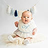 Arnie is registered to the contest to win money with this photo: baby, infant, white_clothing, bib, sitting, fluffy_rug, tassel_garland, indoor, cute, portrait, child, soft_light, studio, happy, curious, head, face, sock, background, decor