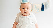Arnie is registered to the contest to win money with this photo: baby, infant, party_hat, white_clothing, sitting, fluffy_rug, celebration, milestone, banner, blue, white_background, cute, portrait, child, indoor, smiling, happy, headwear, sock, person