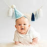 Arnie is registered to the contest to win money with this photo: baby, infant, party_hat, blue_hat, white_clothing, smiling, face, portrait, fluffy_rug, tassel_garland, celebration, half_birthday, cute, happy, indoors, child, young_baby, headwear, soft_lighting, background_blur