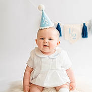 Arnie joined the competition — help win amazing prizes! baby, infant, party_hat, white_dress, socks, fluffy_rug, celebration, half_birthday, smiling, blue, banner, decor, cute, portrait, child, sitting, indoor, happy, milestone, young