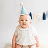baby, infant, party_hat, white_dress, socks, fluffy_rug, celebration, half_birthday, smiling, blue, banner, decor, cute, portrait, child, sitting, indoor, happy, milestone, young