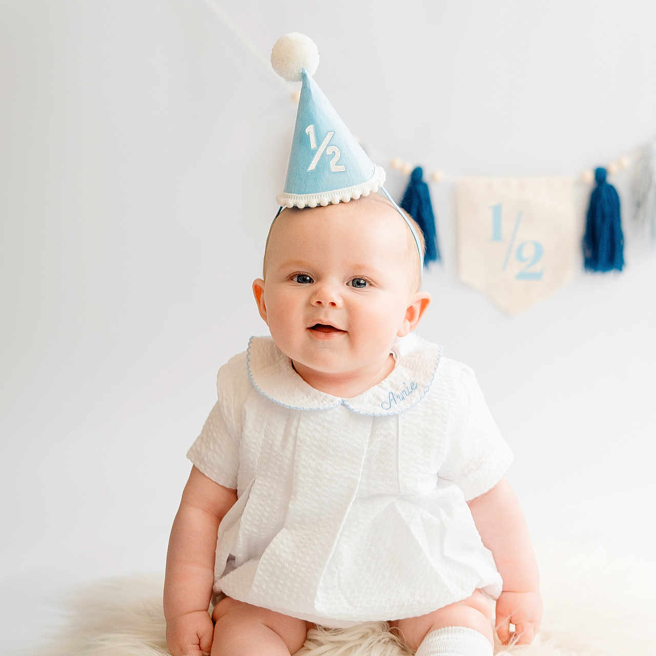 Arnie joined the competition — help win amazing prizes! baby, infant, party_hat, white_dress, socks, fluffy_rug, celebration, half_birthday, smiling, blue, banner, decor, cute, portrait, child, sitting, indoor, happy, milestone, young