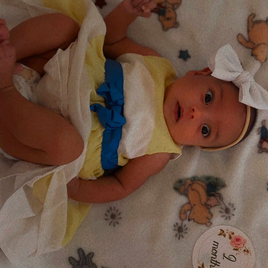 Evelyn joined the competition — help win amazing prizes! baby, infant, child, portrait, lying_down, blanket, teddy_bears, bow, headband, dress, blue_sash, yellow_clothing, white_clothing, wide_eyes, surprised, indoors, bed, soft_blanket, birthday_milestone, two_months
