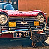 vintage_car, red_car, triumph, classic_car, cat, spotted_cat, outdoor, sunlight, chrome, headlights, license_plate, animal, pet, street, shadow, curious_cat, vehicle, metal, grill, parking