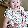 toddler, child, smile, blonde_hair, dress, floral_pattern, sitting, indoor, happy, person, hand, face, cute, young_child, skin, arm, portrait, furniture, light, casual