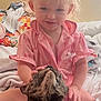 toddler, child, cat, pet, pink_clothing, bed, blanket, indoor, cute, smiling, curly_hair, animal, feline, cozy, playful, young_child, happy, domestic, person, interaction