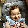 baby, child, curly_hair, tongue_out, smiling, striped_clothing, plush_cushion, indoor, hand, face, happy, person, fabric, patterned_cloth, cute, skin, head, sitting, playful, cozy