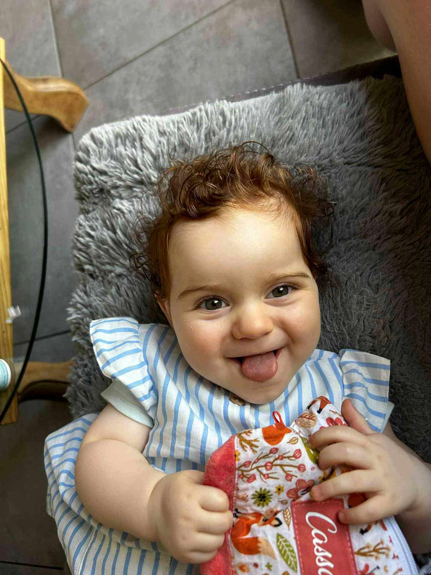 Cassandre a rejoint le concours — aidez-le/la à gagner de superbes lots ! baby, child, curly_hair, tongue_out, smiling, striped_clothing, plush_cushion, indoor, hand, face, happy, person, fabric, patterned_cloth, cute, skin, head, sitting, playful, cozy