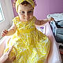 baby_care, baby_item, changing_table, child, clothing, curious_expression, curly_hair, furniture, hand, headband, indoor, nursery, pacifier, person, portrait, purple_wall, sitting, soft_lighting, toddler, yellow_dress