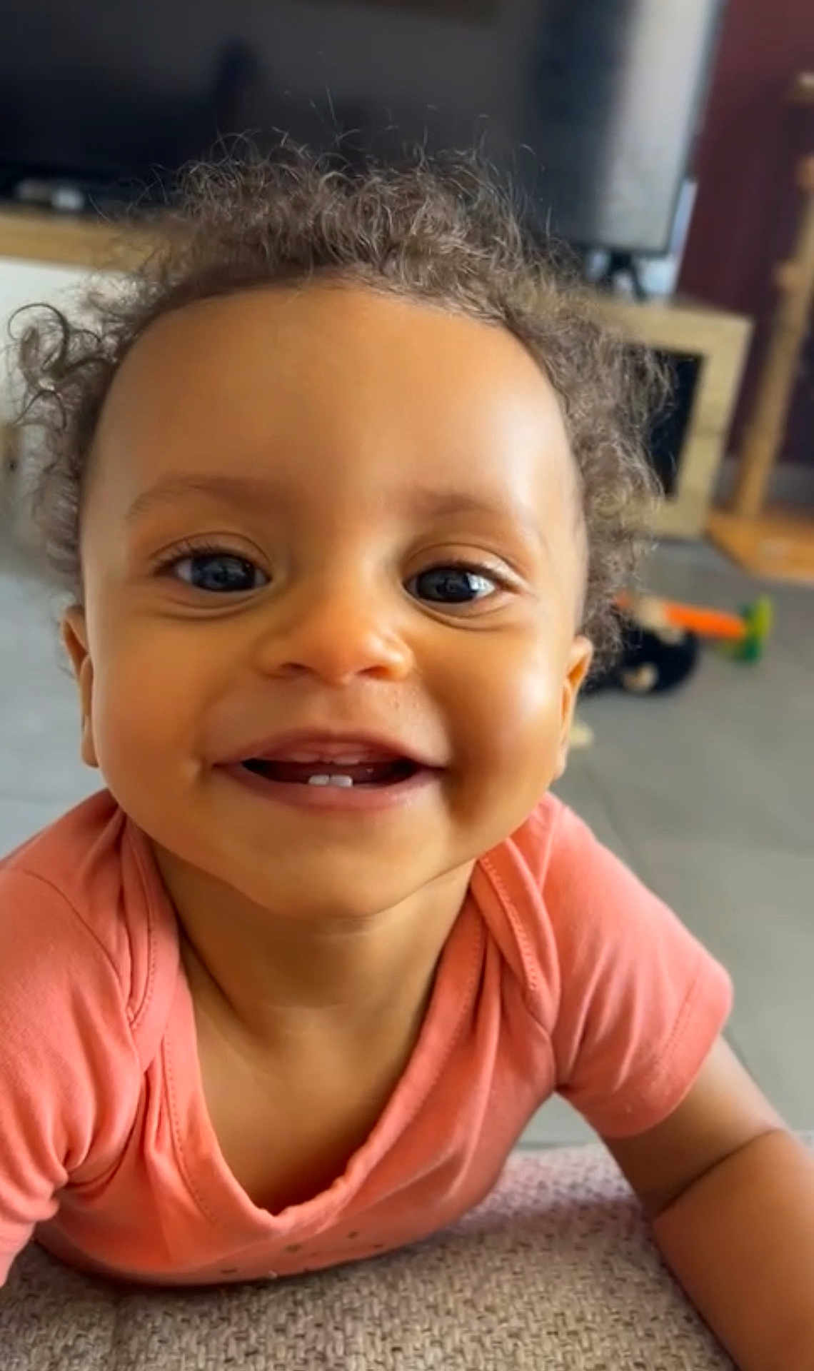 Kélya participe au concours pour gagner de l'argent avec cette photo : child, toddler, smiling, curly_hair, face, teeth, shirt, indoor, living_room, floor, couch, happy, cute, baby, person, portrait, close_up, furniture, home, skin