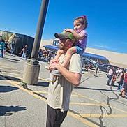 Leah joined the competition — help win amazing prizes! child, man, outdoor, daylight, parking_lot, people, drink, cap, shorts, tshirt, sandals, shadow, blue_sky, curly_hair, holding, walking, crowd, casual, smiling, shoulder_ride