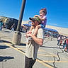 child, man, outdoor, daylight, parking_lot, people, drink, cap, shorts, tshirt, sandals, shadow, blue_sky, curly_hair, holding, walking, crowd, casual, smiling, shoulder_ride