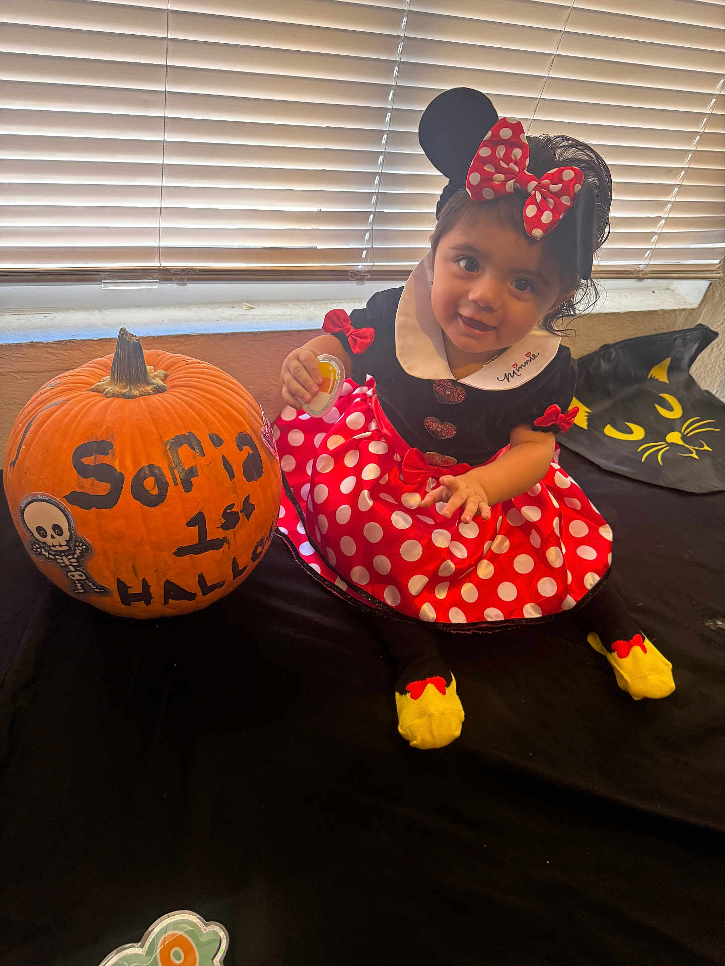 Isa joined the competition — help win amazing prizes! toddler, child, pumpkin, costume, minnie_mouse, polka_dots, red, black, yellow_socks, window, blinds, smile, indoor, table, decoration, halloween, black_cat_design, cute, dress, face