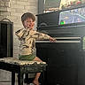 barefoot, bench, brick_wall, child, cozy, curtains, dinosaur_hoodie, easel, flooring, flowers, furniture, green_shorts, home, indoor, music, painting, person, piano, playing, smiling